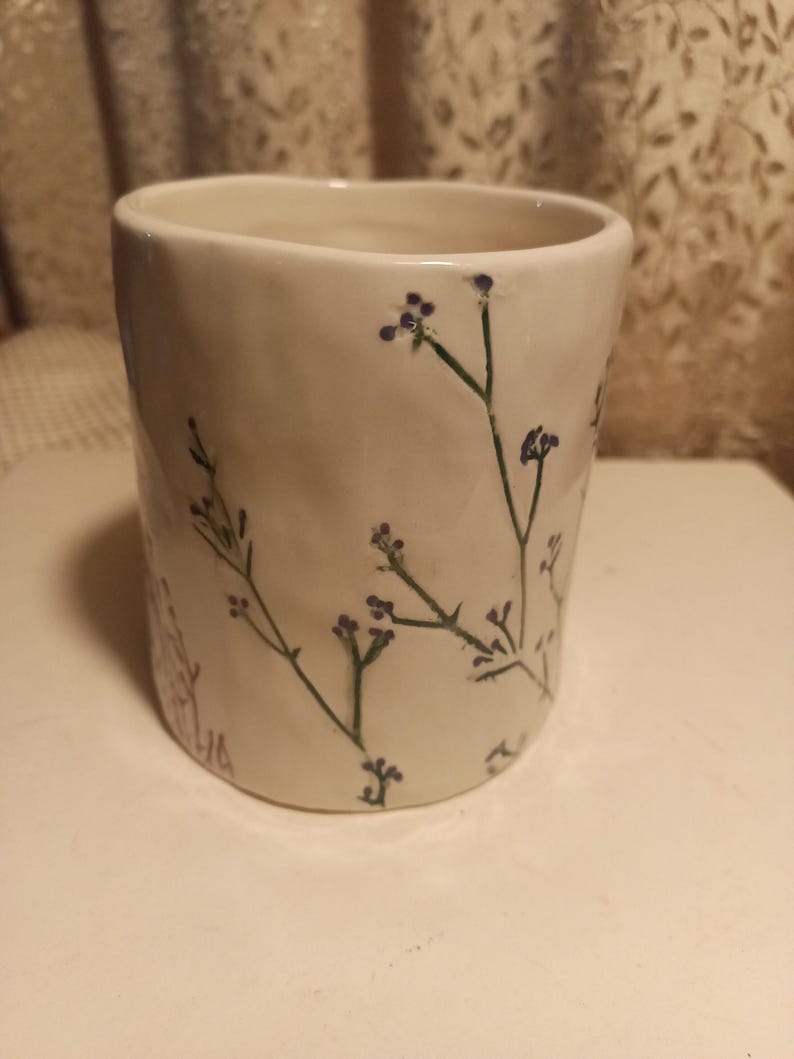 Puede incluir: Una taza de cer&aacute;mica blanca con una superficie ligeramente texturizada. Delicados tallos verdes y peque&ntilde;as flores moradas est&aacute;n pintados a mano en el exterior. La taza tiene forma cil&iacute;ndrica y un borde liso.