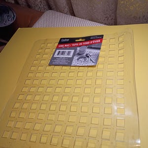 May include: A clear, flexible plastic sink mat featuring a grid of raised square sections. A black product label is attached to the top, displaying the text "Cooking Concepts SINK MAT / TAPIS DE FOND D'&Eacute;VIER" and dimensions of 12 in x 11 in.