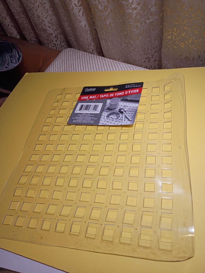 May include: A clear, flexible plastic sink mat featuring a grid of raised square sections. A black product label is attached to the top, displaying the text "Cooking Concepts SINK MAT / TAPIS DE FOND D'&Eacute;VIER" and dimensions of 12 in x 11 in.