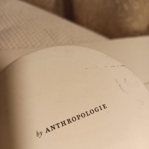 Puede incluir: Objeto redondeado de color blanquecino con el texto "by ANTHROPOLOGIE" impreso en negro. El objeto parece ser parte de un art&iacute;culo m&aacute;s grande, posiblemente una pieza decorativa o un componente de un art&iacute;culo de decoraci&oacute;n del hogar.