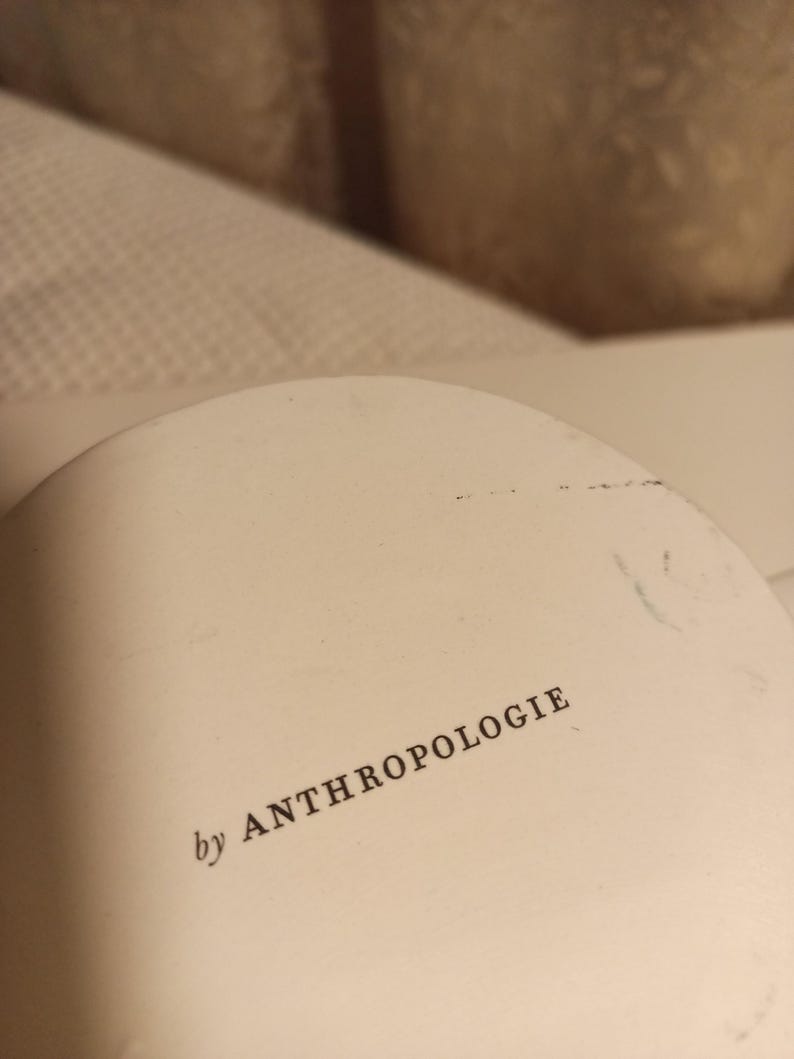 Puede incluir: Objeto redondeado de color blanquecino con el texto "by ANTHROPOLOGIE" impreso en negro. El objeto parece ser parte de un art&iacute;culo m&aacute;s grande, posiblemente una pieza decorativa o un componente de un art&iacute;culo de decoraci&oacute;n del hogar.