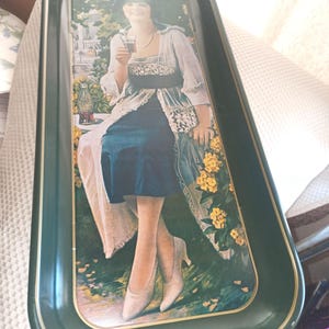 May include: A rectangular dark green metal serving tray featuring a vintage illustration of a person in a white and blue outfit holding a Coca-Cola bottle. The tray has gold-toned trim and the text Drink Coca-Cola printed at the top center.