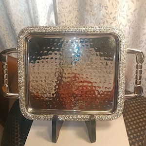 May include: A silver serving tray with a hammered finish and a rhinestone border. The tray has two handles and is sitting on a black stand.