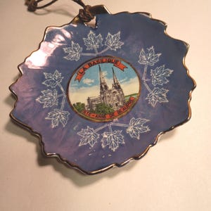 May include: A blue decorative dish shaped like a leaf, with a gold rim and a black string for hanging. The dish features white maple leaf designs and a central image of a church with the text "LA BASILIQUE STE-ANNE DE BEAUPRÉ".
