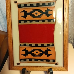 May include: A framed tile with a Southwestern-style design. The tile features a red, orange, black, and white geometric pattern. The frame is made of wood and has a brown finish.