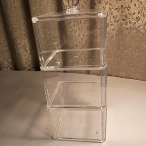 May include: A clear, three-tiered acrylic storage container with a clear, spherical knob on the lid. The container has a rectangular shape with a clear, transparent design. The storage unit is designed to hold small items.