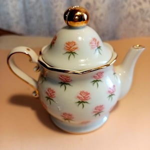 May include: A small, white porcelain teapot with a lid and handle. The teapot is decorated with pink roses and green leaves. The lid has a gold-colored knob. The rim and handle are also trimmed in gold.