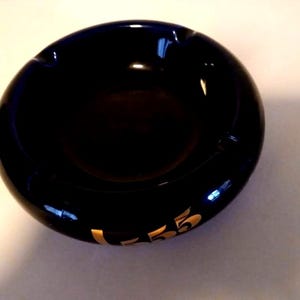 May include: A glossy, black ashtray with a circular design. The ashtray has a curved shape with a recessed area for holding cigarettes. Gold lettering on the side reads "L-75".