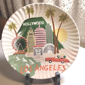 May include: A decorative plate featuring a colorful illustration of Los Angeles landmarks, including the Hollywood sign, palm trees, and a Ferris wheel. The plate has a scalloped edge and the words "HOLLYWOOD" and "LOS ANGELES" are prominently displayed.
