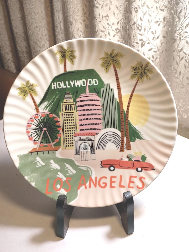 May include: A decorative plate featuring a colorful illustration of Los Angeles landmarks, including the Hollywood sign, palm trees, and a Ferris wheel. The plate has a scalloped edge and the words "HOLLYWOOD" and "LOS ANGELES" are prominently displayed.
