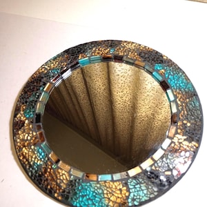 May include: Round mosaic mirror with a black frame. The mosaic border features a mix of teal, gold, and black tiles. The mirror reflects the surrounding environment. Decorative home decor item.