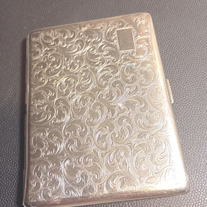 May include: A rectangular silver-toned metal cigarette case featuring an intricate, embossed scrollwork pattern across the entire front surface. A small, smooth, blank rectangular area is positioned in the upper right corner for potential engraving.