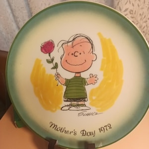 May include: A round ceramic plate with a green border and a yellow and white background. The plate features a cartoon illustration of Charlie Brown holding a red rose. The text "Mother's Day 1972" is printed on the bottom of the plate.