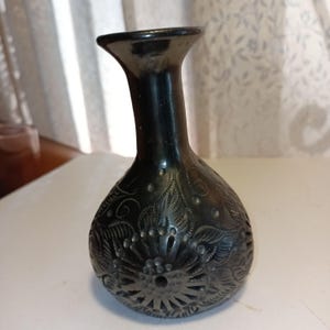 May include: A dark, decorative vase with a flared top and a rounded body. The vase features intricate floral and leaf patterns, along with small, raised dots. The vase is made of a dark, possibly metallic material.