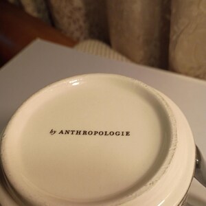 May include: Close-up of a white ceramic mug with the text "by ANTHROPOLOGIE" printed in black. The mug has a patterned design on the side and a silver handle.