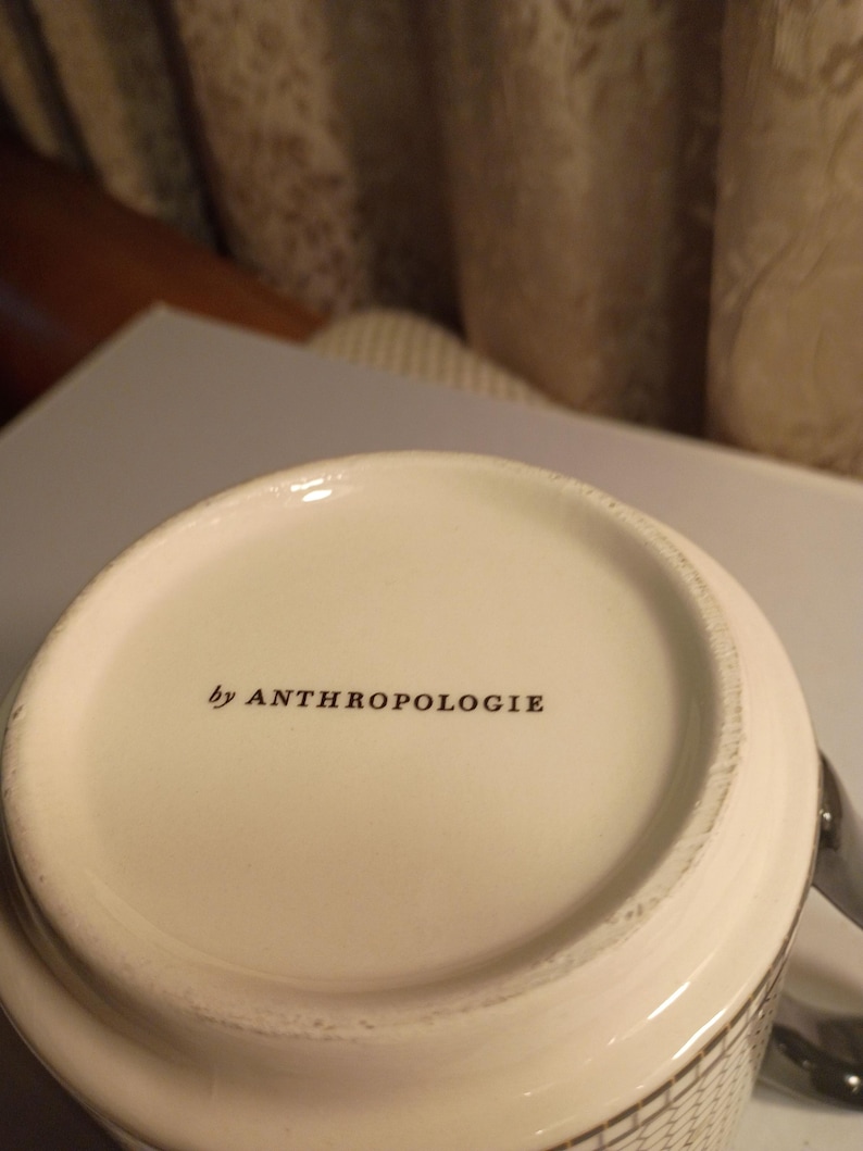 May include: Close-up of a white ceramic mug with the text "by ANTHROPOLOGIE" printed in black. The mug has a patterned design on the side and a silver handle.
