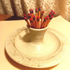 May include: A ceramic match holder with a small, cylindrical cup in the center, surrounded by a wide, shallow dish. The holder is filled with wooden matches, each with a red tip. The ceramic is off-white with a speckled texture.