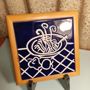 May include: A blue and white ceramic tile with a framed wooden border. The tile features a white illustration of a bowl of noodles with chopsticks on a blue and white checkered background.