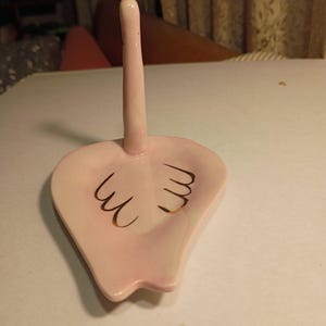 May include: A pink ceramic ring holder with a tall, slender post and a heart-shaped base. The base features gold-colored decorative accents. The ring holder is designed to hold rings and other small jewelry items.