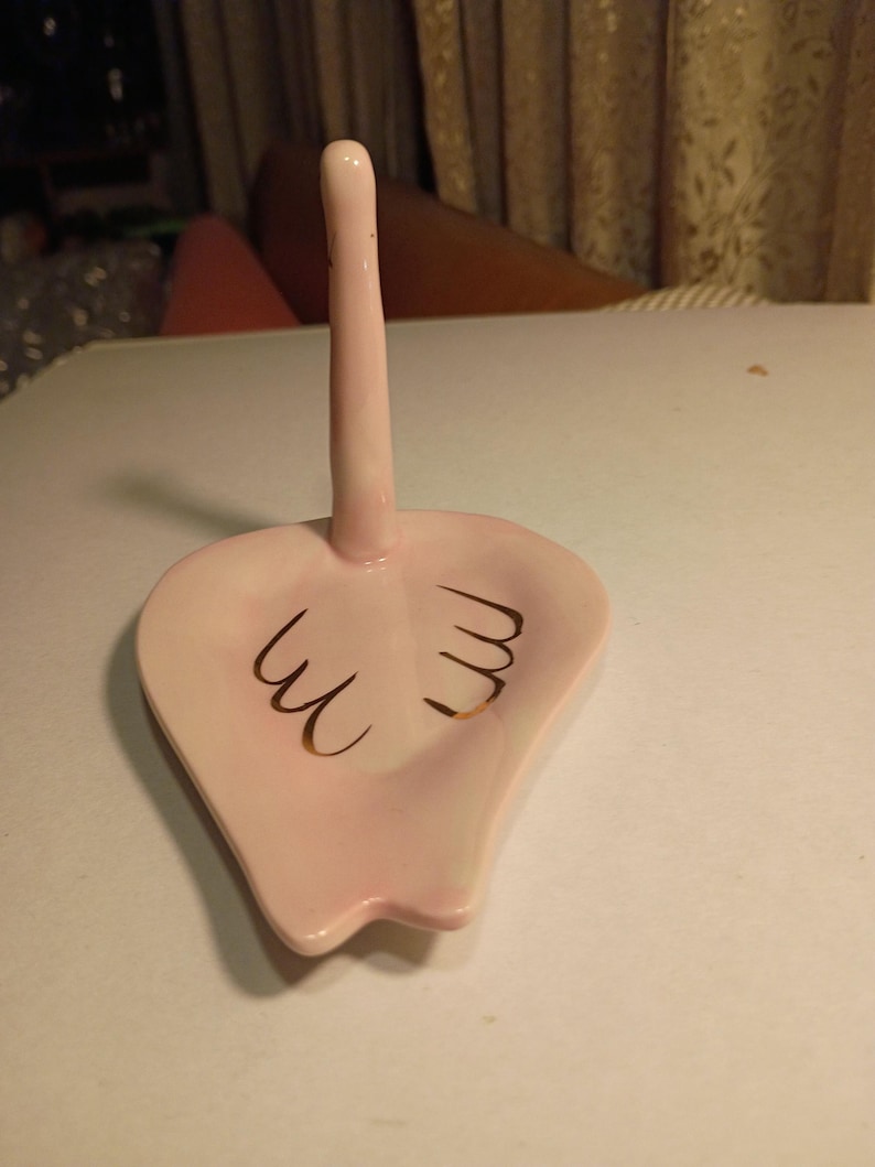 May include: A pink ceramic ring holder with a tall, slender post and a heart-shaped base. The base features gold-colored decorative accents. The ring holder is designed to hold rings and other small jewelry items.