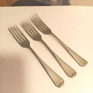 May include: Three antique silver forks with heart-shaped tips. The forks are arranged diagonally across a white surface. Each fork has four tines and a long, slender handle. The silver has a slightly tarnished appearance, suggesting age.
