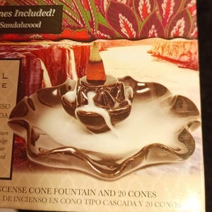 May include: A brown ceramic incense cone fountain with a white smoke plume rising from the top. The fountain is on a black and brown wavy plate. The text on the box reads "Incense Cone Fountain and 20 Cones".