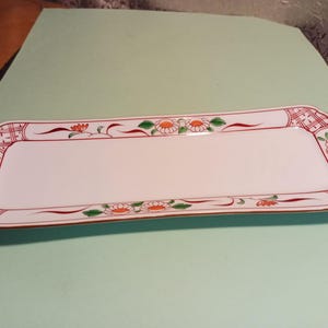 May include: A white rectangular serving dish with a red, green, and orange floral design. The dish has a raised rim and a slightly curved edge.