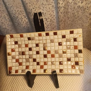 May include: Rectangular mosaic tile tray with a black metal stand. The tray features a pattern of small, square tiles in white, brown, and gold. The stand has a curved design, providing support for the tray. A decorative item for the home.