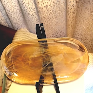 May include: An oval-shaped, amber-colored glass dish with a black stand. The dish has a translucent appearance with swirling patterns. The stand is made of black material and supports the dish. The background features a patterned curtain.