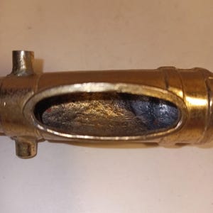 May include: A brass-colored, cylindrical object with an oval opening. The object has two small, cylindrical protrusions and a series of horizontal bands. The interior of the oval opening appears dark.