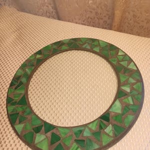 May include: A round, mosaic-style decorative frame. The frame is made of a dark brown material and is decorated with various shades of green triangular glass pieces. The frame is circular with a hollow center.