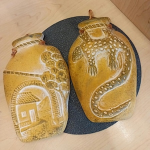 May include: Two ceramic wall hanging planters, one with a brown and white design of a house and tree, the other with a brown and white design of a lizard.