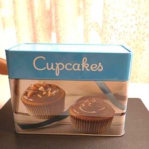 May include: A rectangular light blue metal storage tin featuring the word Cupcakes in white script on the lid. The front panel displays a photographic print of two chocolate frosted cupcakes with brown paper liners and a dark ribbon accent.