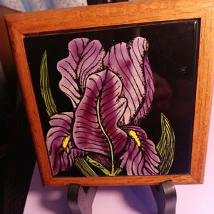 May include: A framed tile with a purple iris flower painted on a black background. The tile is in a wooden frame and is on a black easel.