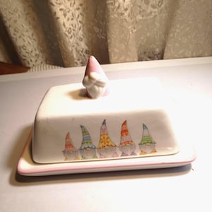 May include: A white ceramic butter dish with a rectangular base and a matching lid. The lid features a pink gnome-shaped handle and a row of five colorful gnomes printed on the side. The base has a thin pink rim around the edge.