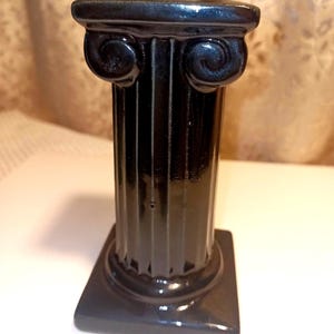 May include: A black, glossy, miniature Ionic column with a square base. The column features fluted sides and decorative scrollwork at the top. The overall design is a classic, architectural style, suitable for display.