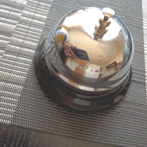 May include: A polished chrome service bell with a rounded dome and a small vertical plunger button at the top. The metallic surface features a reflective finish, and the base is constructed from a solid black material, providing a stable foundation.