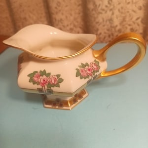 May include: A white porcelain creamer with a gold handle and floral design. The creamer has a hexagonal base and is decorated with pink roses and green leaves.