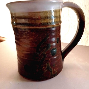 May include: Handmade ceramic mug with a large handle. The mug features a dark brown base with dark green and black accents. The rim is light purple with a band of brown and tan. The mug is approximately 5 inches tall.
