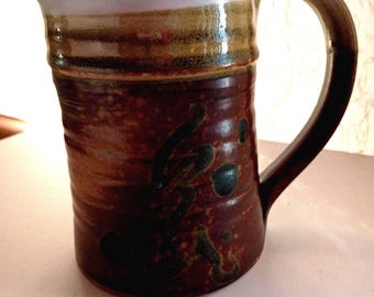 May include: Handmade ceramic mug with a large handle. The mug features a dark brown base with dark green and black accents. The rim is light purple with a band of brown and tan. The mug is approximately 5 inches tall.