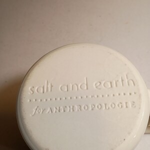 Peut inclure: Base de tasse en c&eacute;ramique blanc cass&eacute; avec texte en relief "salt and earth" au-dessus de "for ANTHROPOLOGIE". La tasse a une forme arrondie et une petite anse est visible sur le c&ocirc;t&eacute; droit.