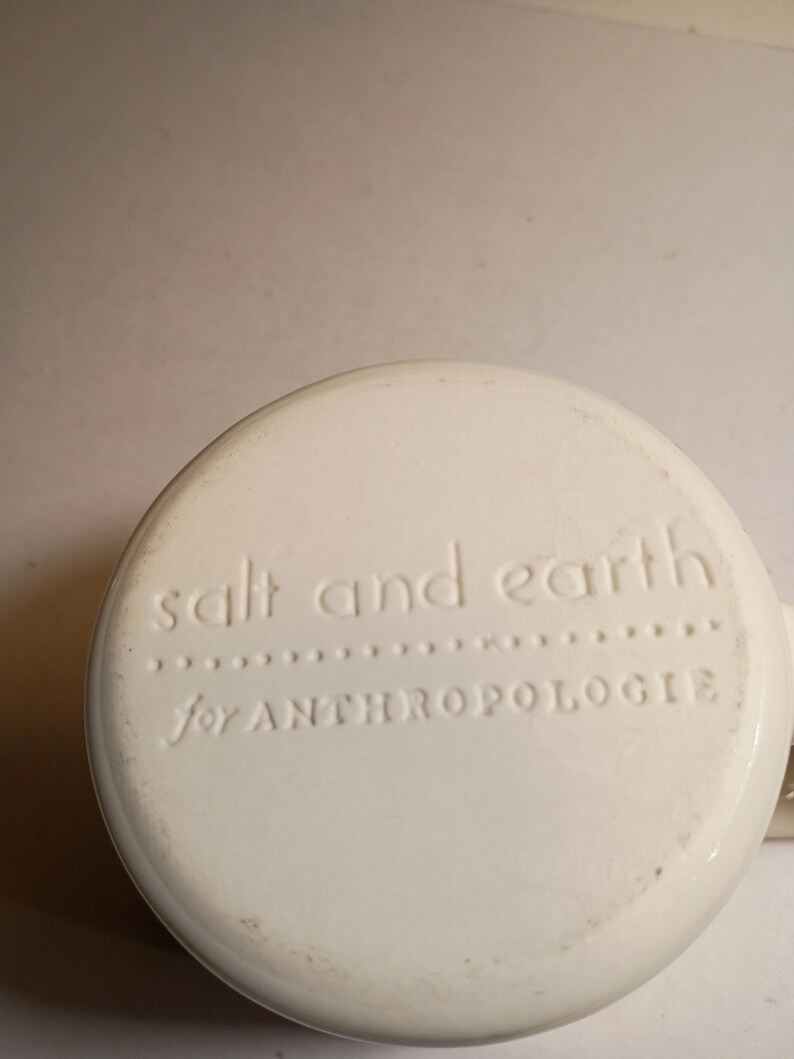 Peut inclure: Base de tasse en c&eacute;ramique blanc cass&eacute; avec texte en relief "salt and earth" au-dessus de "for ANTHROPOLOGIE". La tasse a une forme arrondie et une petite anse est visible sur le c&ocirc;t&eacute; droit.