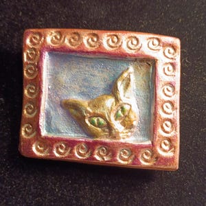 May include: A rectangular, handmade brooch featuring a cat's head within a frame. The cat is gold with green eyes, set against a light blue background. The frame is a reddish-brown with a spiral design.