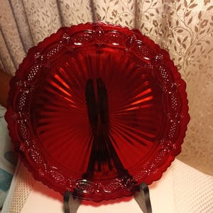 May include: A vibrant red glass serving plate with a decorative edge and a radial pattern. The plate is supported by a black stand, showcasing its intricate design. This vintage piece adds a touch of elegance to any table setting.