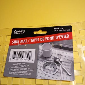 May include: A clear, rectangular sink mat with a grid of square cutouts, featuring a black and red product label that states 12 in x 11 in. The label displays a barcode, the text Cooking Concepts, and a photo of the mat in a kitchen sink.
