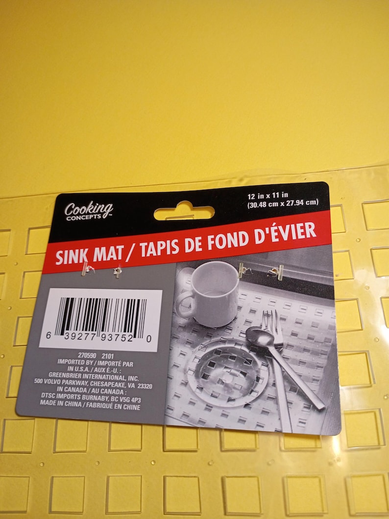 May include: A clear, rectangular sink mat with a grid of square cutouts, featuring a black and red product label that states 12 in x 11 in. The label displays a barcode, the text Cooking Concepts, and a photo of the mat in a kitchen sink.