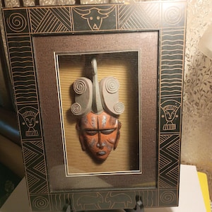 May include: A framed, three-dimensional African mask sculpture. The mask is reddish-brown with carved details, set against a textured gold background. The dark brown frame features geometric and animal-inspired designs. The piece is displayed on a black stand.