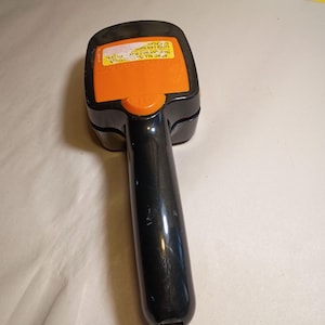 May include: A black handheld grooming tool with an orange rectangular top section. The orange panel features a yellow sticker with black text that reads READ ALL INSTRUCTIONS. The tool has a long, smooth black handle and a rounded head design.