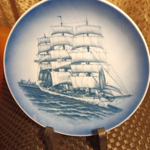 May include: A blue and white ceramic plate with a detailed illustration of a sailing ship. The plate is on a stand and has a decorative border.