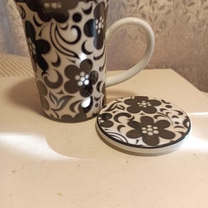 May include: A tall ceramic mug with a matching lid. The mug and lid feature a repeating floral pattern in black and white. The mug has a white handle and a flared top.